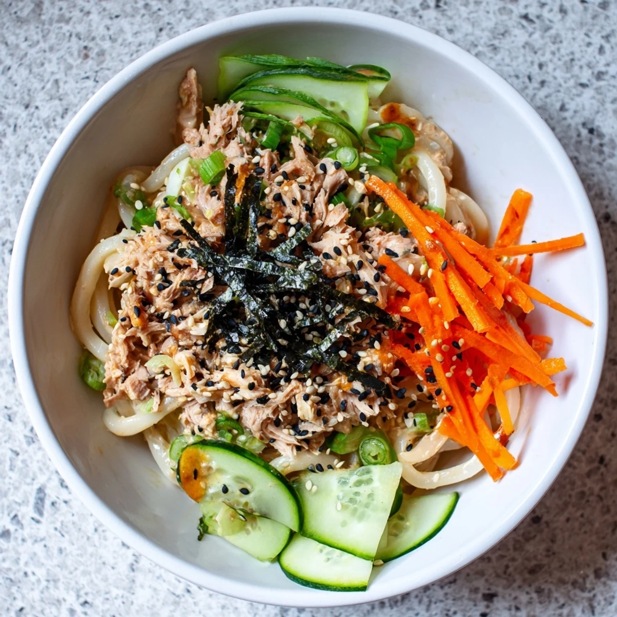 Steaming bowl of spicy tuna udon noodles topped with fresh vegetables, sesame seeds, and nori strips